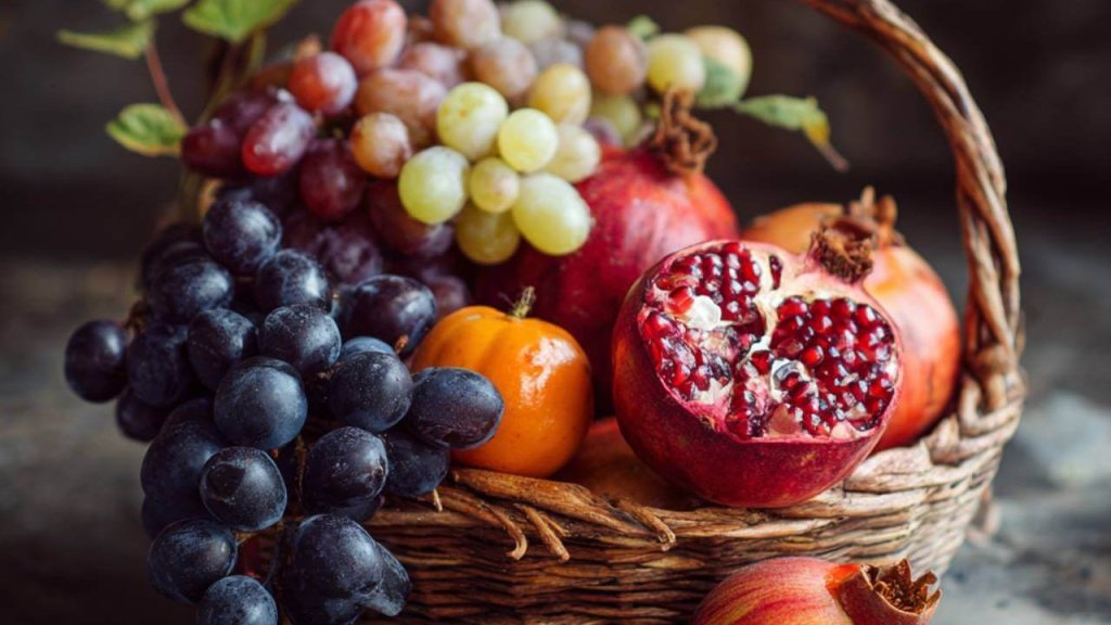 Frutas de otoño variadas — uvas, granadas, mandarinas y manzanas — en una cesta sobre fondo rústico.