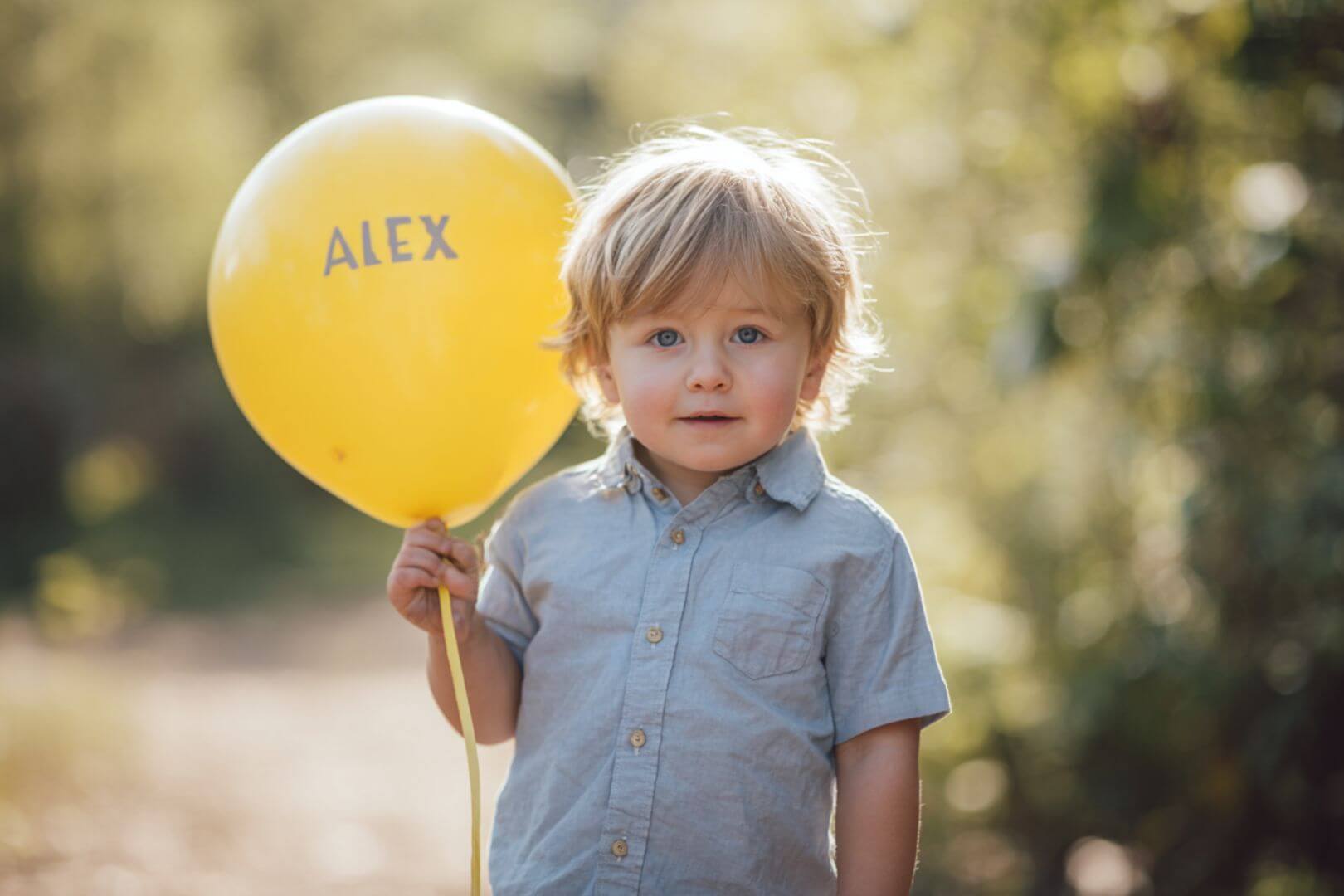 Niño pequeño sosteniendo un globo amarillo con el nombre ÁLEX al aire libre.