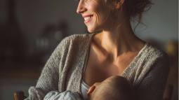 Mujer joven amamantando a su bebé en casa, con luz cálida y expresión sonriente, transmitiendo calma y ternura.