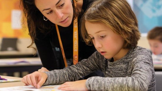 Niño de primaria leyendo un libro en clase con la ayuda de su profesora.