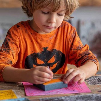 Niño con camiseta naranja de Halloween pintando dibujos con esponja sobre una mesa decorada con calabazas y materiales de manualidades.