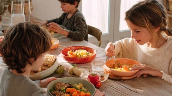 Tres niños sentados a la mesa comiendo platos saludables con verduras, frutas y cereales integrales en una casa luminosa.