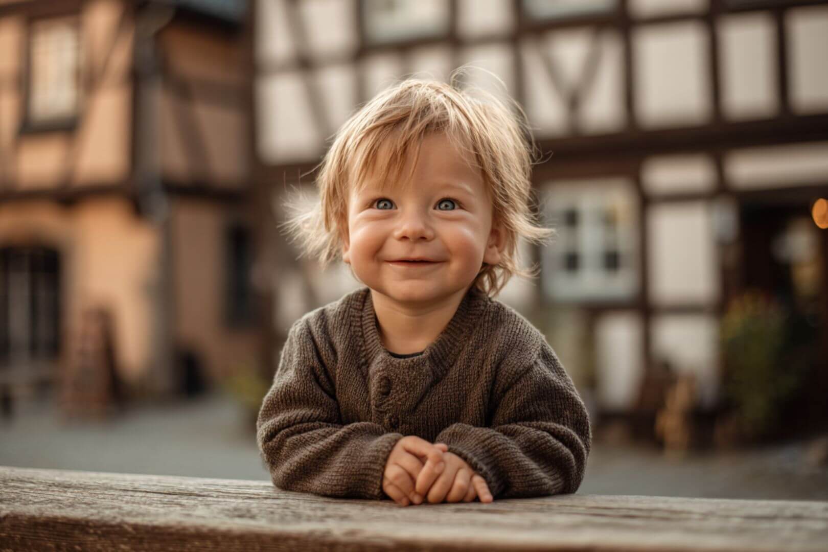 Bebé rubio envuelto en suéter marrón sonriendo frente a casas de entramado típicas de Alemania.