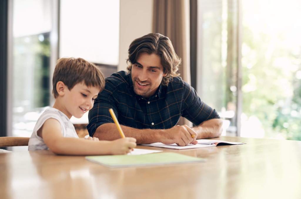 Padre con media melena y camisa de cuadros e hijo con pelo corto haciendo los deberes en el salón de casa, sobre una mesa de madera