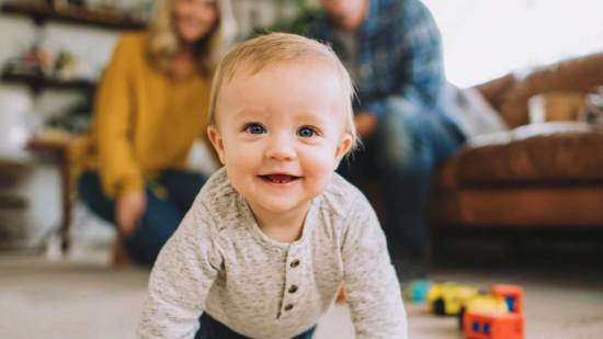 Bebé gateando y sonriendo, con sus padres al fondo en un ambiente familiar acogedor.