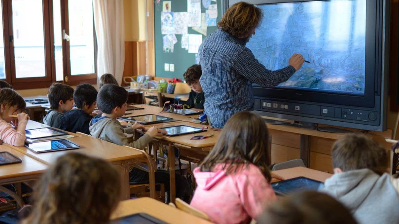 Aula de primaria en España con un profesor enseñando en pizarra digital y niños usando tablets en sus mesas.
