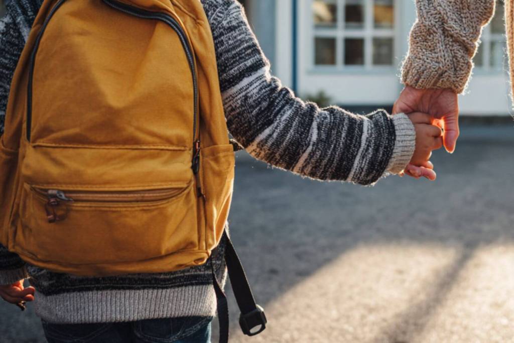 Detalle de un niño con mochila amarilla cogido de la mano de un adulto, camino al colegio durante la vuelta a clase.