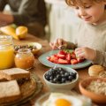Niño eligiendo qué comer en el desayuno entre frutas, yogur y pan integral, acompañado por su madre en una cocina luminosa.