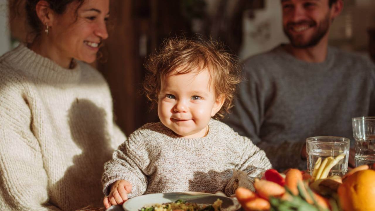 Bebé sonriente sentado a la mesa con sus padres, rodeado de frutas y verduras frescas durante la comida familiar.