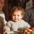 Bebé sonriente sentado a la mesa con sus padres, rodeado de frutas y verduras frescas durante la comida familiar.