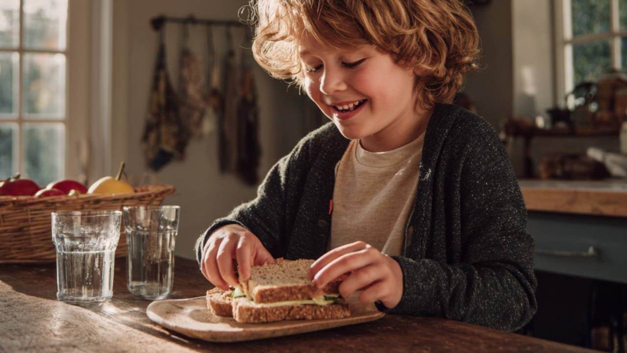 Niño merendando un sándwich saludable en casa con fruta y agua.