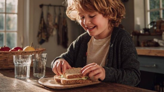 Niño merendando un sándwich saludable en casa con fruta y agua.