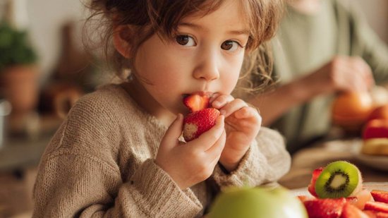 Niño probando distintas frutas con curiosidad mientras su madre le anima en una cocina luminosa.