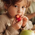 Niño probando distintas frutas con curiosidad mientras su madre le anima en una cocina luminosa.
